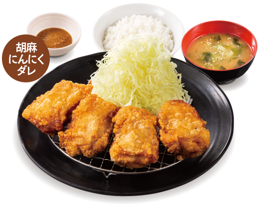 Four large pieces of fried chicken on a black plate. Shredded fresh cabbage is also served on the side. White Rice and miso soup. Sesame garlic sauce is available on a separate plate.